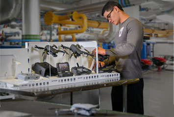 A student works on a detailed model of a ship, surrounded by tools and equipment in a workshop setting.