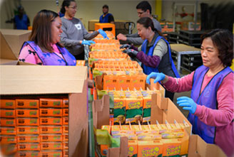 Volunteers pack food boxes in a warehouse, contributing to community support and outreach efforts.