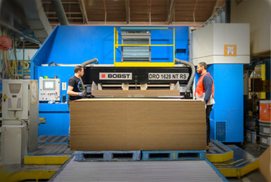 Two workers operate a BOBST DRO 1628 NT RS machine, processing large sheets of cardboard in a manufacturing facility.