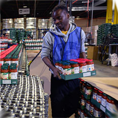 A worker in a warehouse carefully carries boxes of jars, contributing to food distribution efforts.