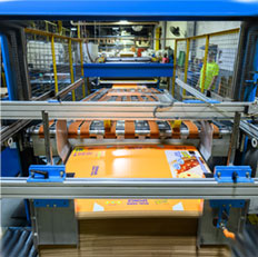 Automated printing machinery processes vibrant orange sheets in a busy production facility.