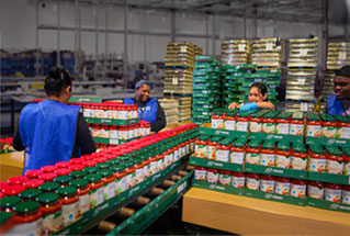 Volunteers organize jars of sauce in a warehouse, showcasing community efforts in food distribution.