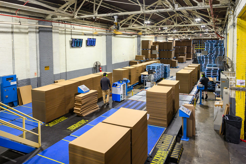 A busy warehouse with workers handling stacked cardboard boxes and pallets, showcasing the company's operational environment.