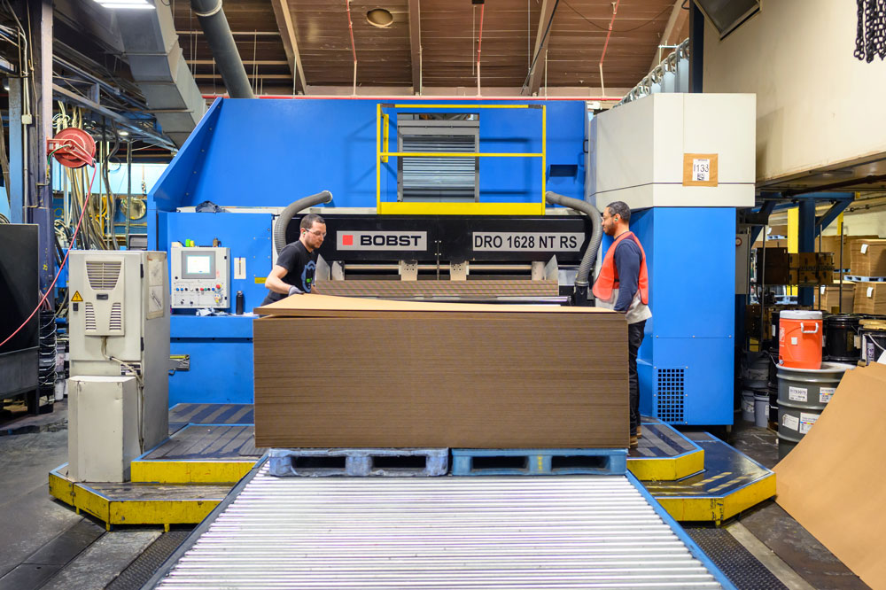 Two workers operate a large Bobst machine in a manufacturing facility, processing sheets of cardboard.