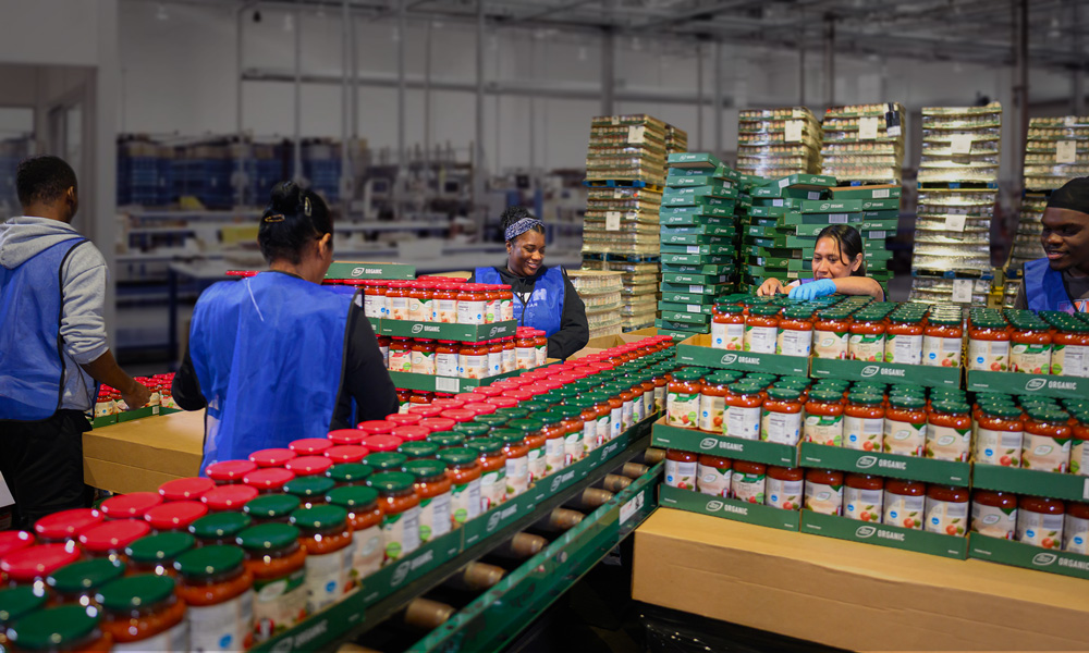 Workers in blue vests efficiently pack jars of sauce in a busy co-packing fulfillment warehouse.