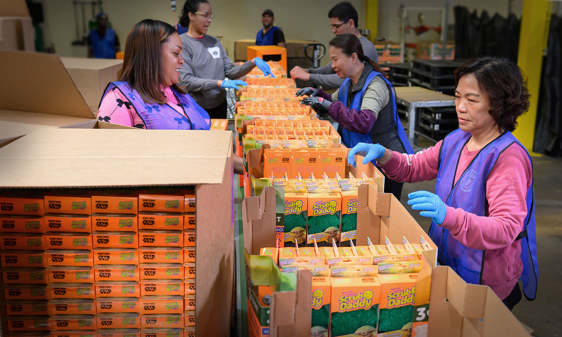 Volunteers package Scrub Daddy products in a warehouse, showcasing teamwork in industrial packaging efforts.