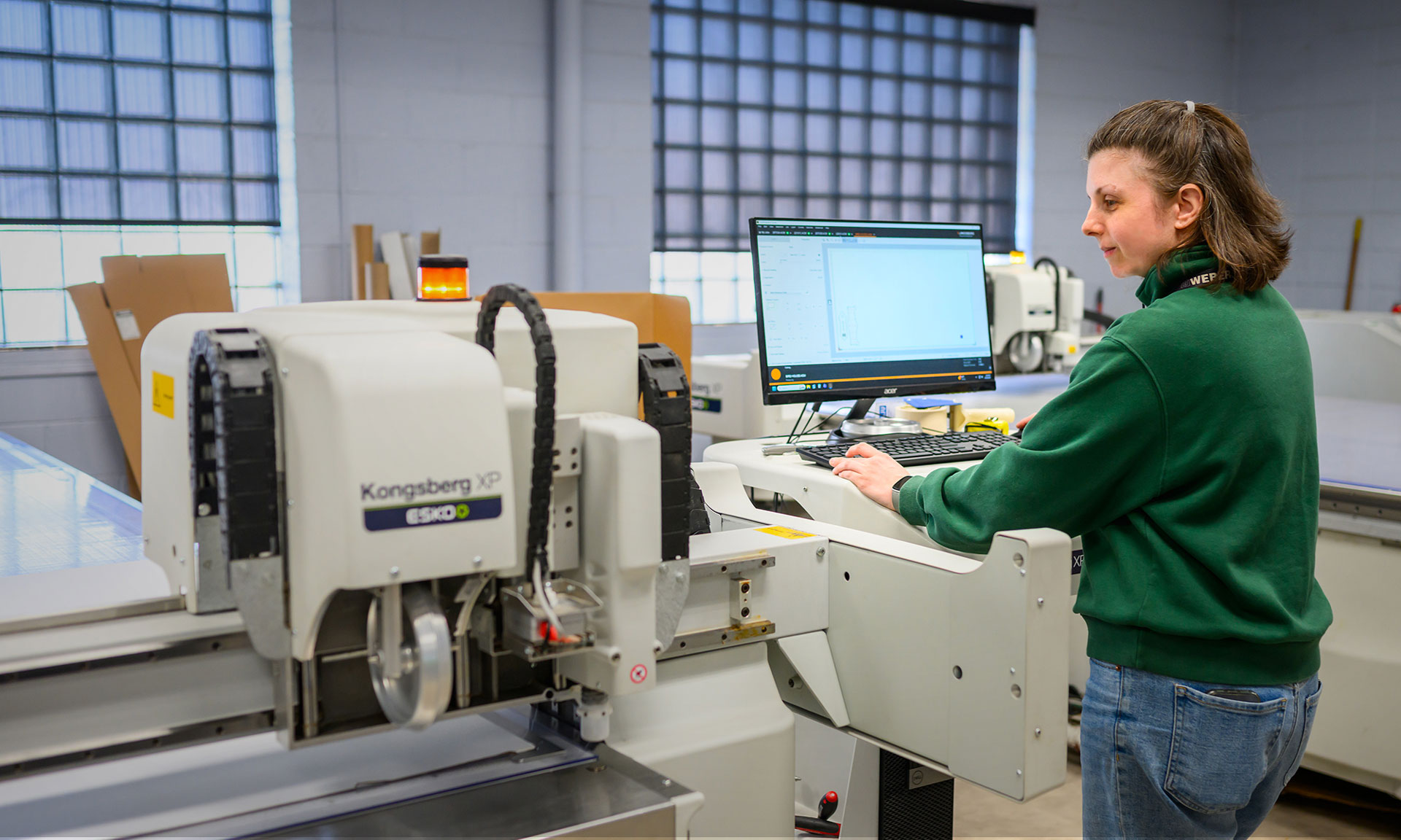 A worker operates a Kongsberg XP cutting machine while using a computer in an industrial packaging facility.