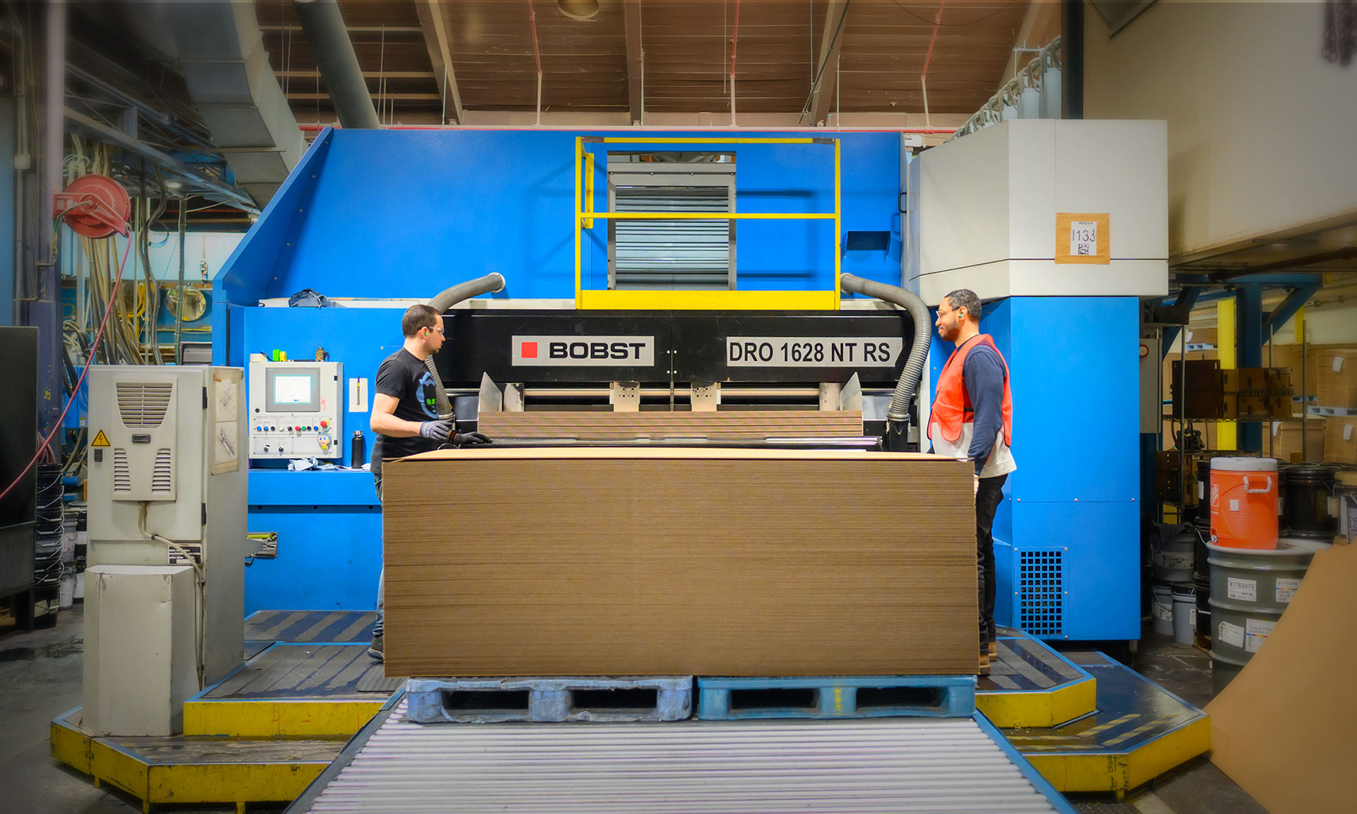Two workers operate a BOBST industrial machine for packaging cardboard in a manufacturing facility.