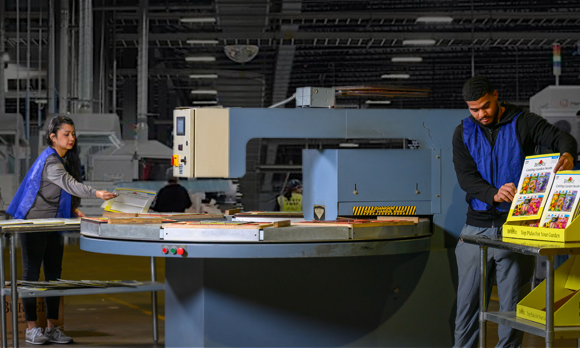 Workers operate machinery in an industrial packaging facility, handling printed materials and preparing them for distribution.